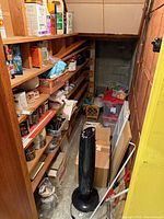 Narrow storage area with shelves of paint supplies, tower fan in foreground