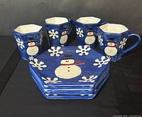 Front view of four mugs and stack of four hexagonal plates showing snowman motif