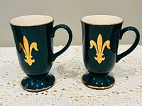 Front view of both dark green footed mugs with gold fleur-de-lis