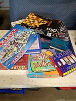 Stack of various board game boxes including Monopoly Here And Now, Yahtzee Deluxe, Chatter Matters, Solitaire, Chess, Pictionary, Spider Wars, Trivial Pursuit cards
