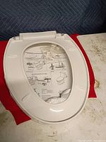 Top view of white elongated toilet seat still wrapped with instruction sheet and hardware visible