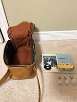 Open tan vinyl camera case next to manuals, viewfinder, flash bulbs and film