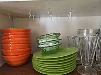 Stack of orange bowls, green plates, green dessert bowls, clear parfait glasses on shelf