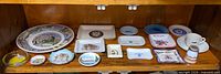 Shelf holding large New Mexico plate, multiple small ceramic souvenir trays, teacup and saucer, clear glass ashtray, and shot glass