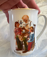 Mug showing toymaker scene, gold rim visible, Norman Rockwell signature at base