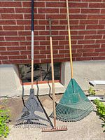 Three rakes standing against brick wall: black plastic fan rake, metal bow rake, green plastic fan rake