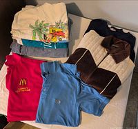 Stacked garments showing white graphic sweatshirt, denim sweatshirt, velour pullover, pink McDonald's polo and two light-blue polos