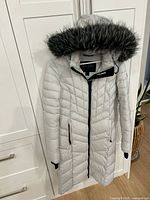 Full view of light grey Pajar long parka hanging, hood with faux fur, shows length and quilting