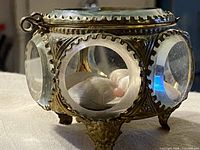 Side view of round brass and beveled glass casket on feet, satin cushion visible