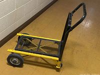 Side view of yellow steel platform truck dolly showing black handle, mesh deck, two large wheels and front caster