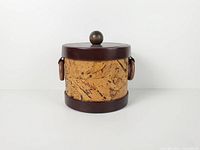 front view of cylindrical cork ice bucket with faux leather trim, side ring handles and lid with teak knob