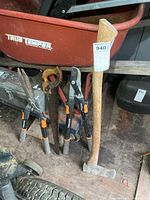 Group of tools leaning against wheelbarrow: axe, dual-blade saw, three Fiskars extendable tools