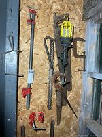 Group of tools hung on wall: pipe clamp, saws, trouble light, pry bar, C-clamp