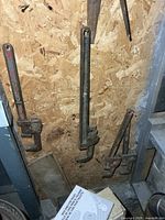 Three pipe wrenches hanging on plywood wall plus partial saw visible