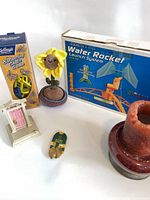 Group shot showing all six items including boxed water rocket, balancing game, crochet sunflower, miniature door, earrings, red candle