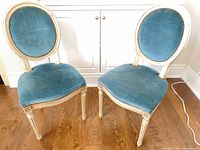 Front view of both chairs showing teal velvet seats and backs, cream distressed frames