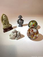 Five items laid out: two glass paperweights, soapstone figure, stone animal, pewter bust