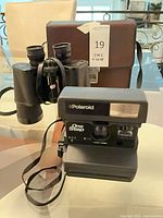 Polaroid OneStep camera, Bushnell 10x50 binoculars, brown case overview