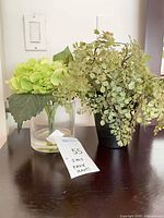 Both faux plants side by side: hydrangea in glass vase, fern in black pot