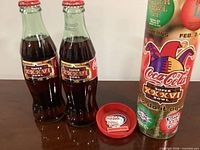 Two commemorative Coca-Cola bottles, cardboard tube, red lid with pin
