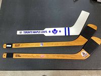 Three miniature hockey sticks laid on table; Detroit Red Wings and New York Rangers wood-finish sticks with printed signatures, Toronto Maple Leafs white goalie stick