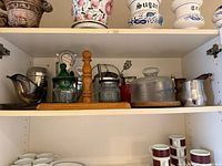 Cabinet shelf showing mixed kitchenware items including canisters, glass jars, pepper grinder, moka pot, cups and saucers