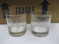 Two glasses showing etched “A” design and heavy bases in front of shipping box