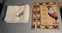 Stack of four cream embroidered placemats beside two rooster tapestry placemats