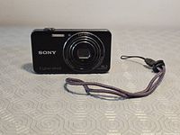 Front view of Sony DSC-WX50 with lens, branding, wrist strap visible