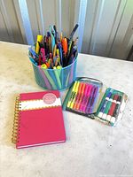 Group shot of pen holder with assorted writing instruments, notebook, two unopened packs