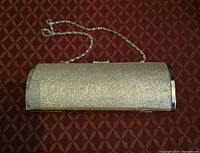 Front view of silver glitter hard-shell clutch with chain strap displayed on patterned surface