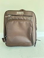 Front view of brown Roots backpack showing exterior zip pockets and top handle