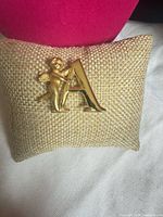 Front view of gold-tone cherub and letter A brooch on burlap cushion