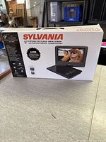 Front of sealed box showing Sylvania 9" portable DVD player image and key features.