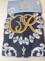Front view of folded navy cotton tea towel with gold and white King Charles III cypher under packaging header