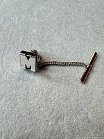 Front view of square tie tack showing raised letter M, chain and T-bar