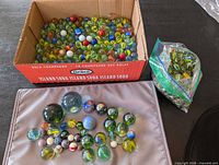 Cardboard box filled with multicolour glass marbles, resealable bag of marbles beside it, large shooters displayed