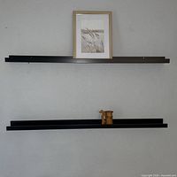 Two dark narrow floating shelves mounted on wall, upper shelf holds framed print, lower shelf holds small wooden decor piece