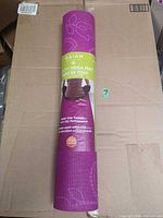 Full view of rolled purple Gaiam Bloom yoga mat in packaging on cardboard background