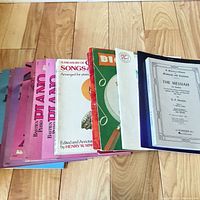 Stack of assorted piano instruction and sheet music books spread on wooden floor