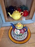Group shot showing platter, multicolour teapot and five solid-colour teapots stored in cabinet