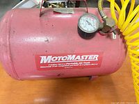 Front view of red MotoMaster 5 gal air tank with branding, gauge, yellow coiled hose