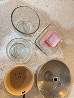 Top-down view of all six kitchen items: aluminum angel food pan, ceramic colander, two glass bowls, glass baking dish, pink silicone cup