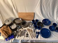Full lot layout showing enamel plates, bowls, mugs, colander, pots, utensils, cutting board, cloth bags