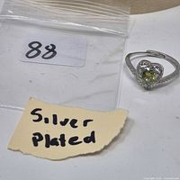 Ring front view showing green heart stone, clear halo, silver tone band, handwritten 'Silver Plated' note