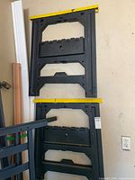 Two folded black plastic sawhorses with yellow top rails hanging on wall