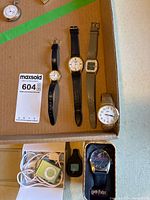 Overview showing four wristwatches on cardboard, metal Timex at right, Quidditch watch in tin, iPod Shuffle with earbuds and charger, Fitbit tracker with clip