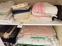 Shelf with assorted folded sheets, pink striped coverlet, king duvet cover on lower shelf, woven throws visible