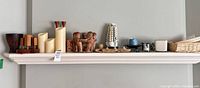 Full shelf showing candles, wood holders, carved sculpture, white column, natural wood tray, assorted small holders, wicker basket