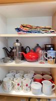 Cabinet shelf with teapots, French press, tea infuser, mugs and quilt